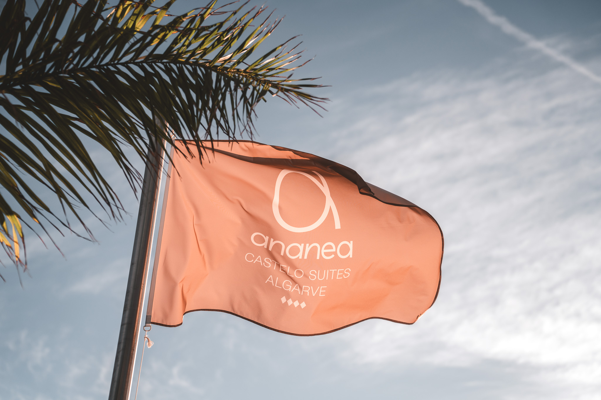 „Orangefarbene Fahne mit Logo und Schriftzug ‚ananea Castelo Suites Algarve‘ weht im Wind, eingerahmt von Palmenblättern vor blauem Himmel.“