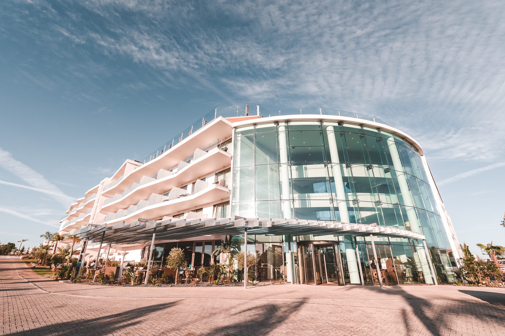 „Modernes Hotelgebäude mit eleganter Glasfassade, Balkonzimmern und mediterraner Architektur unter blauem Himmel – stilvoller Eingangsbereich für Gäste.“