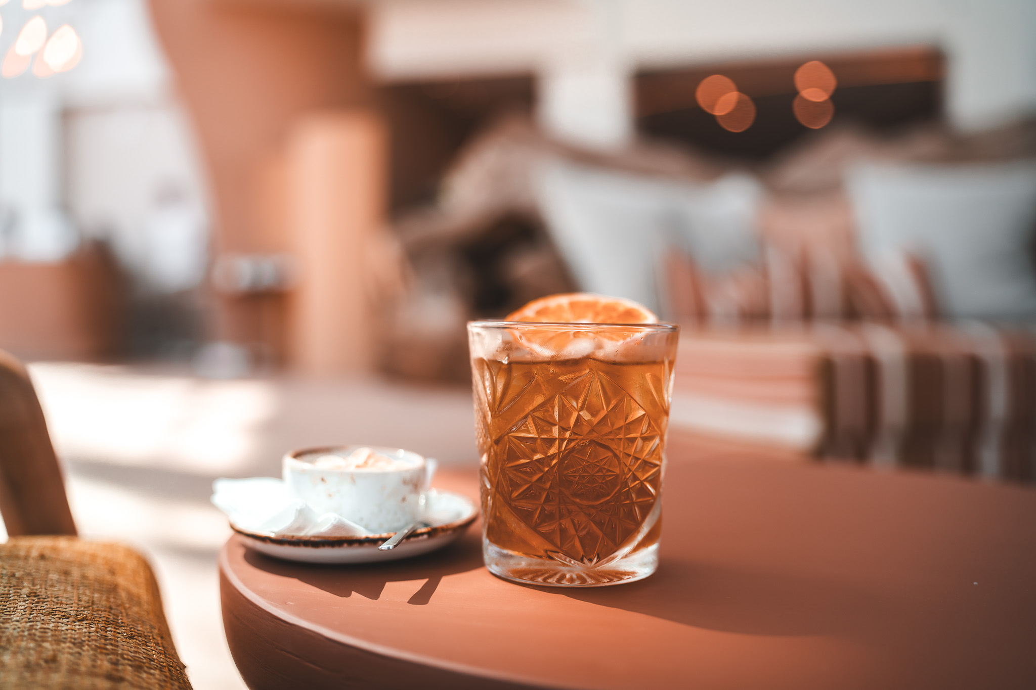 „Stilvoll servierter Cocktail mit Orangenscheibe und eine Tasse Cappuccino auf einem Tisch im modernen Lounge-Bereich.“