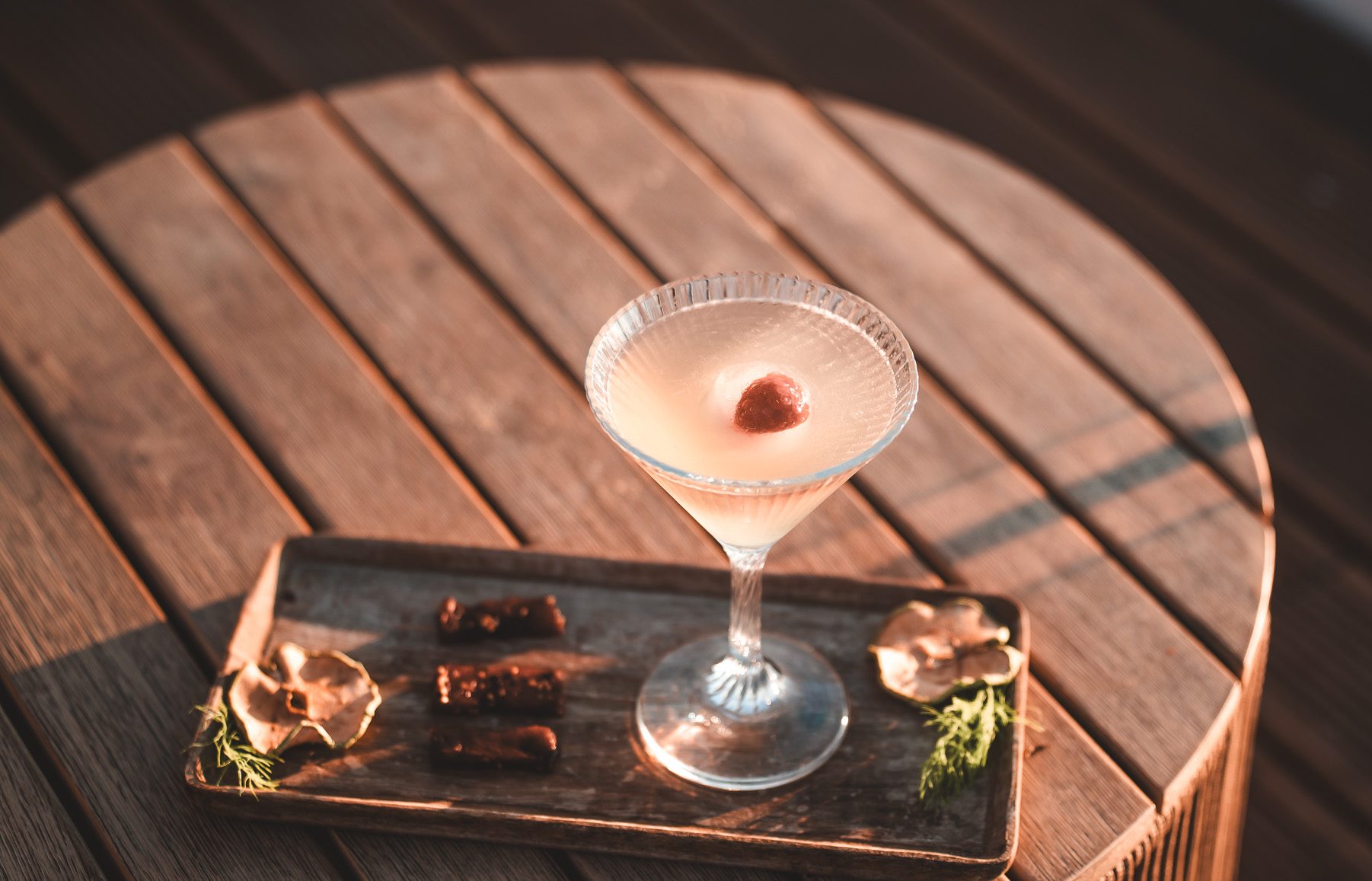 „Eleganter Cocktail im Martiniglas mit Garnitur, serviert auf einem Holztisch mit kleiner Snackplatte in stilvollem Ambiente.“