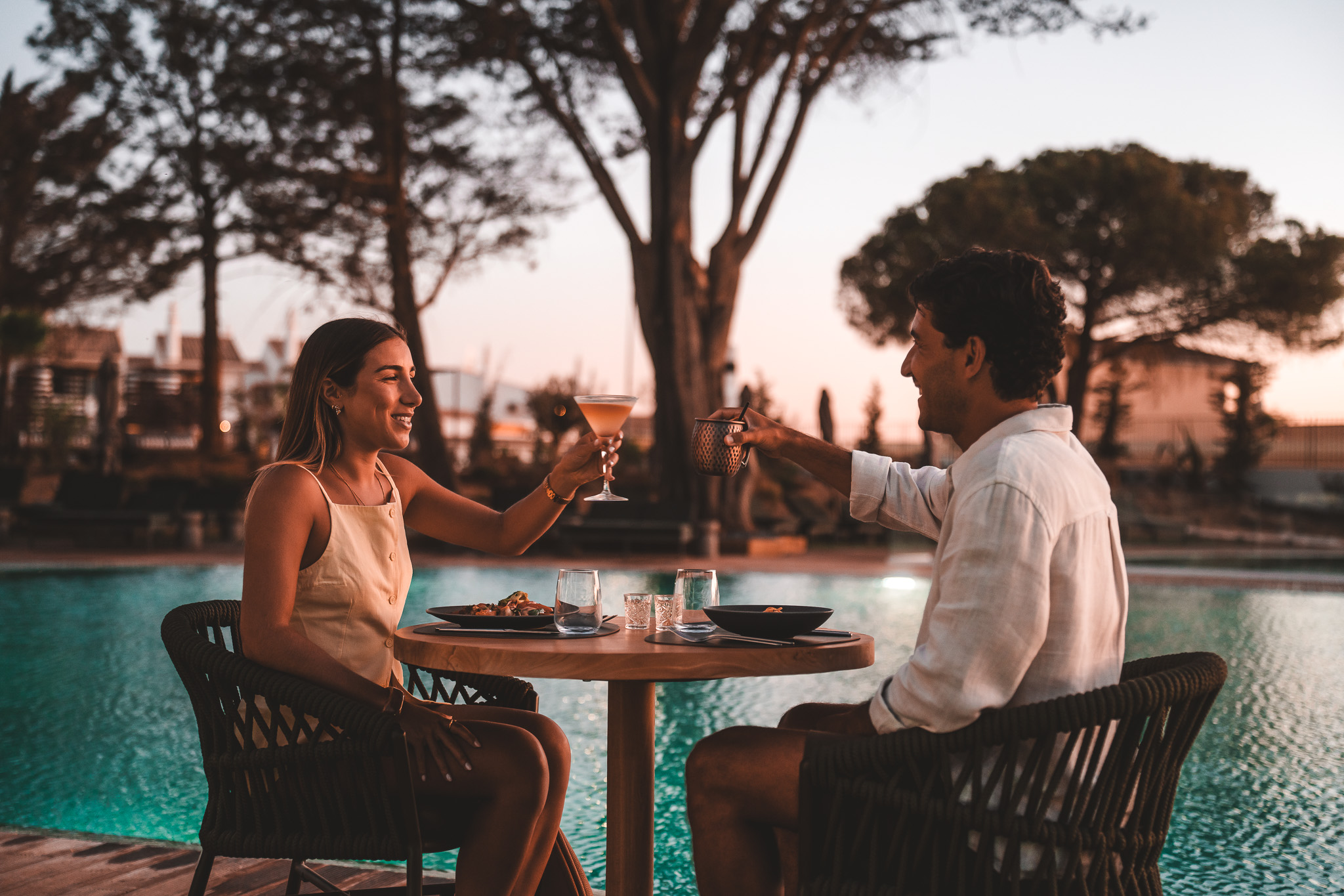 „Romantisches Abendessen am Pool bei Sonnenuntergang – ein Paar stößt an und genießt die besondere Atmosphäre unter freiem Himmel.“