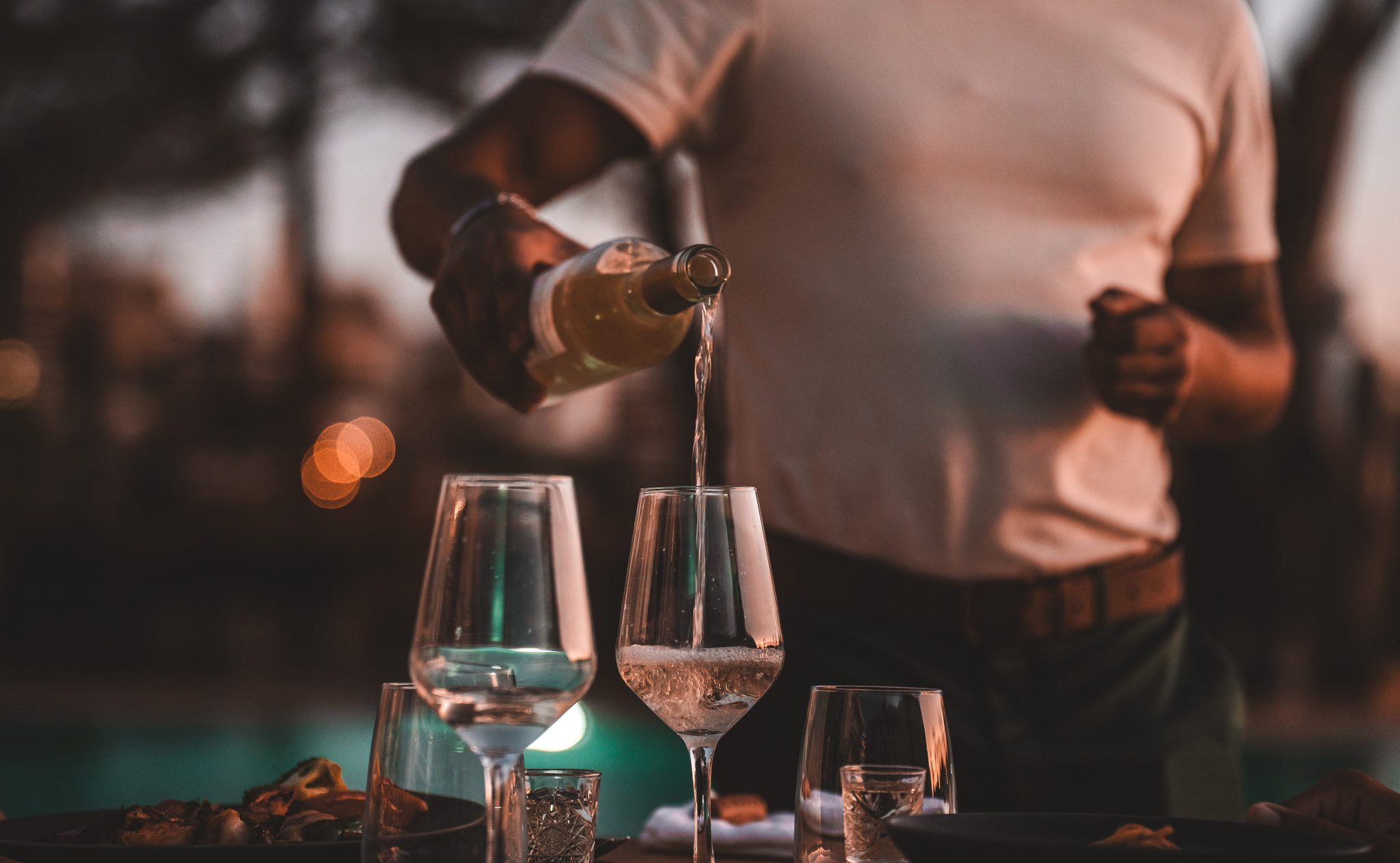 „Kellner schenkt Weißwein in elegante Gläser bei stimmungsvoller Abendatmosphäre – Genussmoment am Pool bei Sonnenuntergang.“
