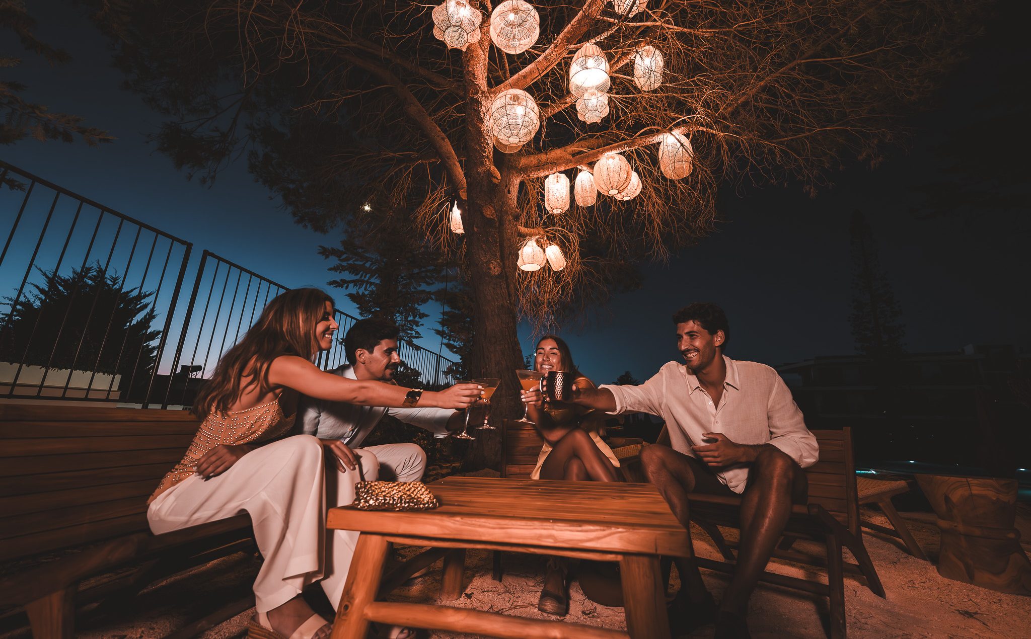 „Gruppe von Freunden stößt abends bei Drinks unter einem dekorativ beleuchteten Baum im Außenbereich an – stimmungsvolle Sommeratmosphäre.“