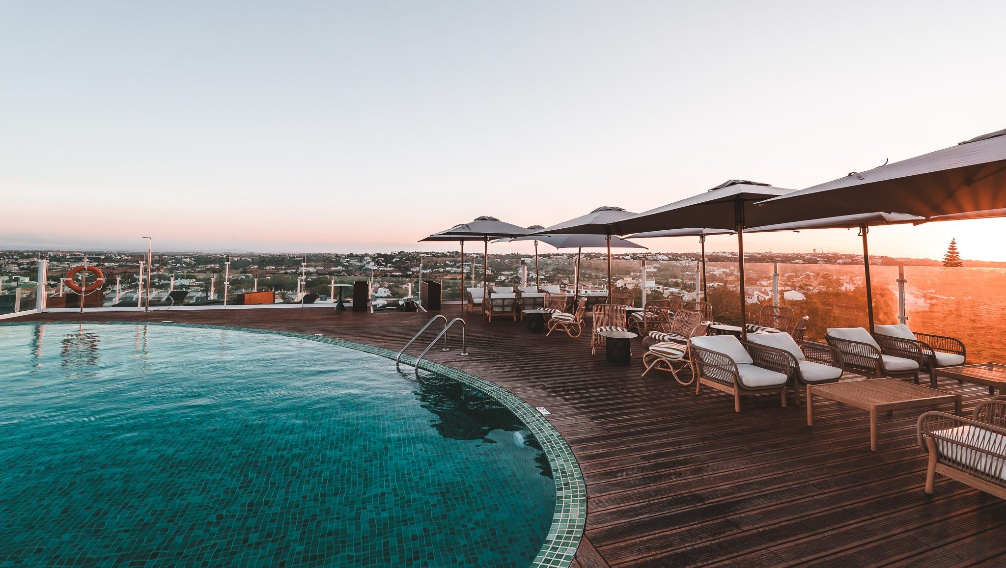 „Rooftop-Pool mit stilvollen Loungemöbeln und Sonnenschirmen, Blick über die Dächer der Stadt im warmen Licht des Sonnenuntergangs – perfekte Atmosphäre zum Entspannen.“