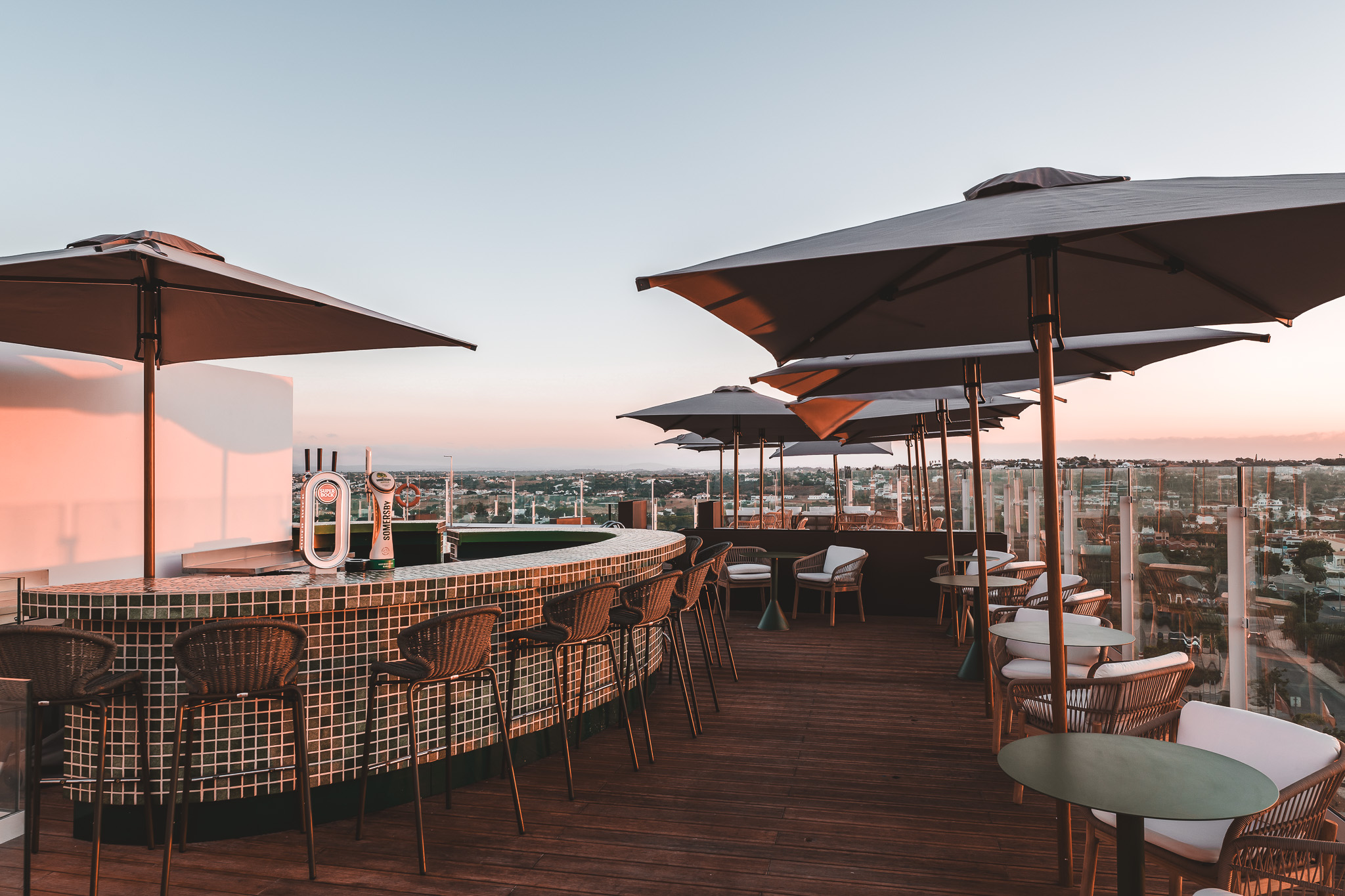 „Stylische Rooftop-Bar mit Theke, Barhockern und gemütlichen Sitzbereichen unter Sonnenschirmen, mit Panoramablick über die Stadt im Abendlicht.“