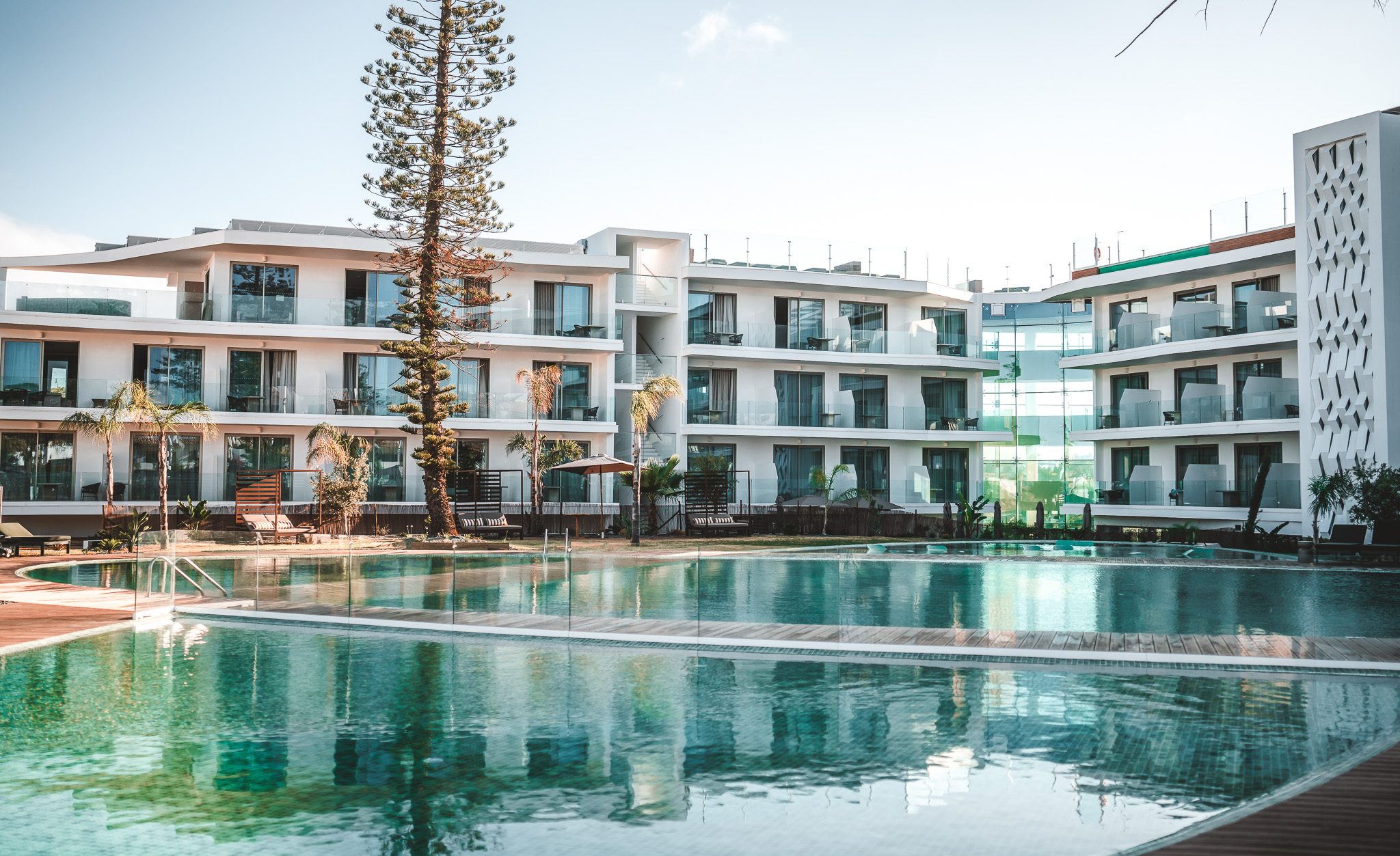 „Moderne Hotelanlage mit Balkonzimmern und großzügiger Poollandschaft, umgeben von Palmen und Glasfassaden im Sonnenlicht.“