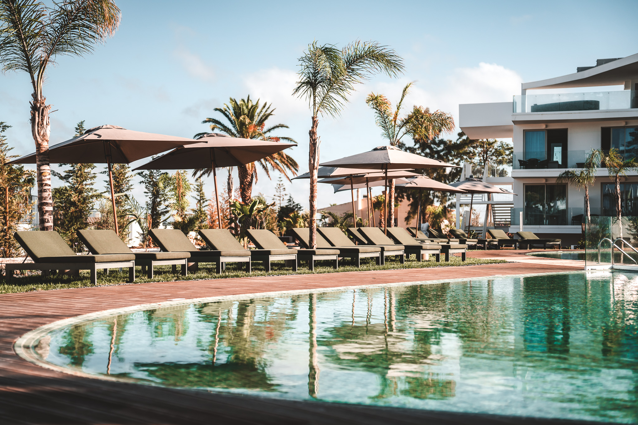 „Pool mit Sonnendeck, Liegen und Sonnenschirmen, umgeben von Palmen, vor moderner Hotelanlage – entspannte Atmosphäre für sonnige Urlaubstage.“