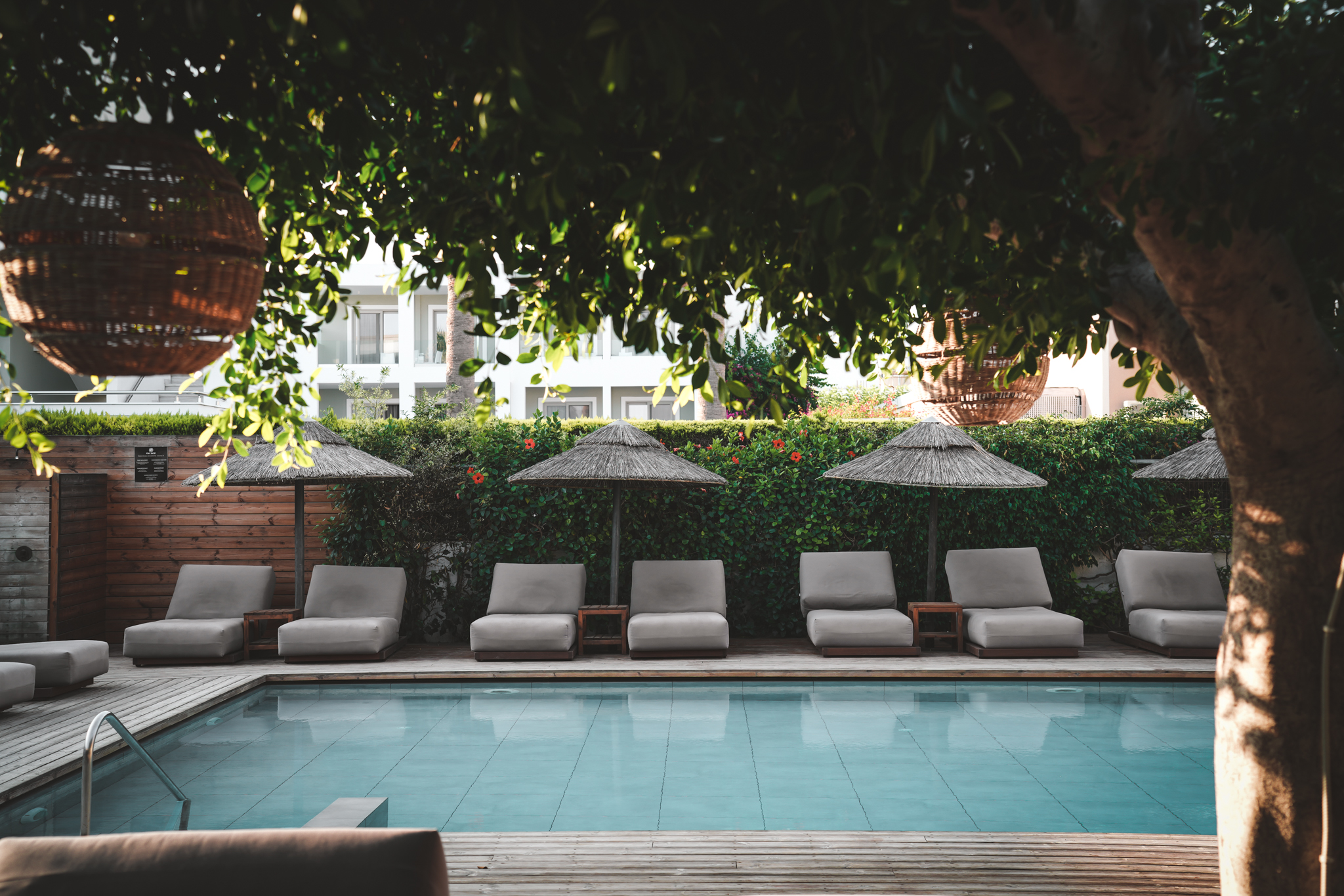 Ruhiger Hotelpool mit gepolsterten Sonnenliegen und Strohdach-Sonnenschirmen, umrahmt von grüner Hecke und Bäumen, die angenehmen Schatten spenden.