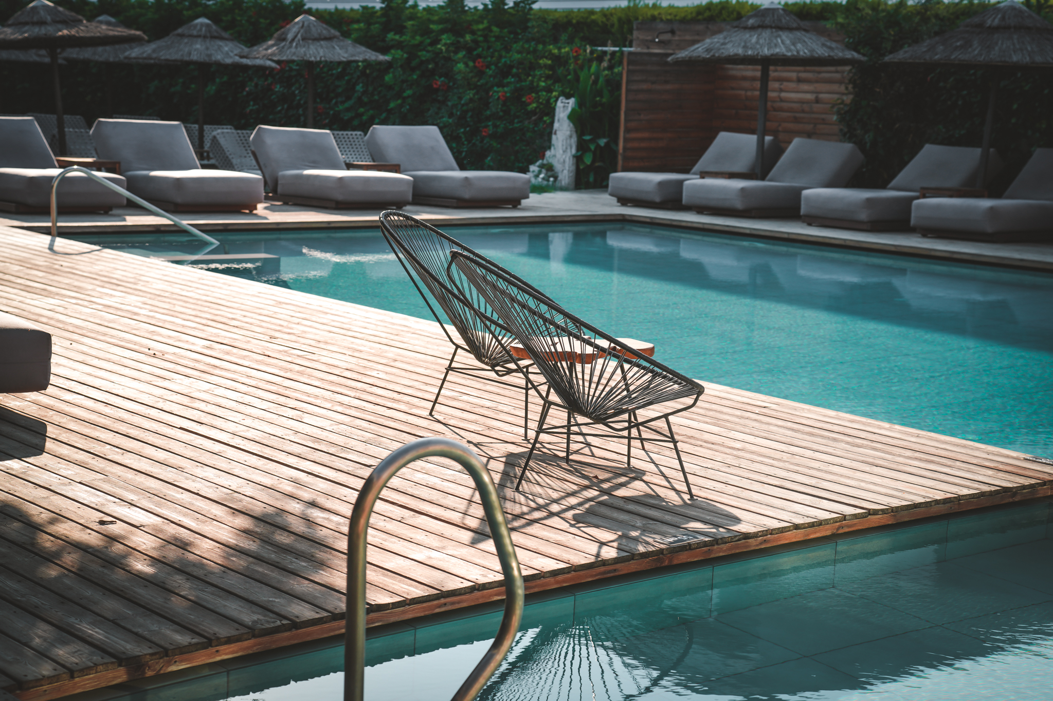 Moderner Drahtstuhl auf einer Holzterrasse am Pool, umgeben von Sonnenliegen und Sonnenschirmen in einer entspannten Urlaubsatmosphäre.