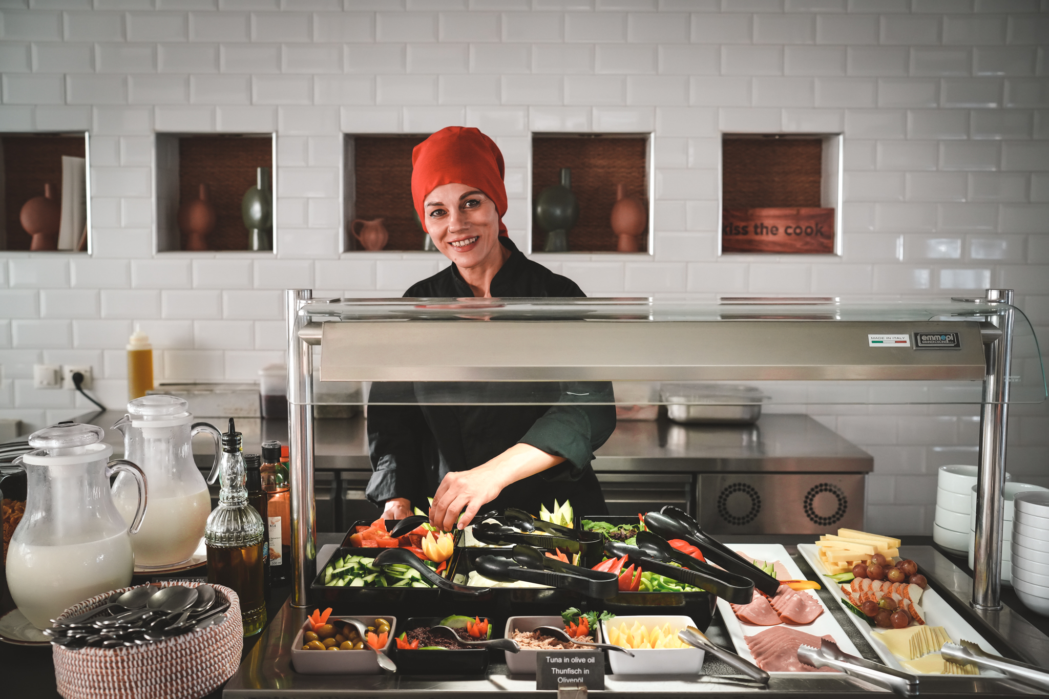 Freundliche Köchin mit roter Kopfbedeckung richtet frisches Gemüse und Aufschnitt an einem Buffet in modernem Restaurant mit weiß gefliester Wand an.