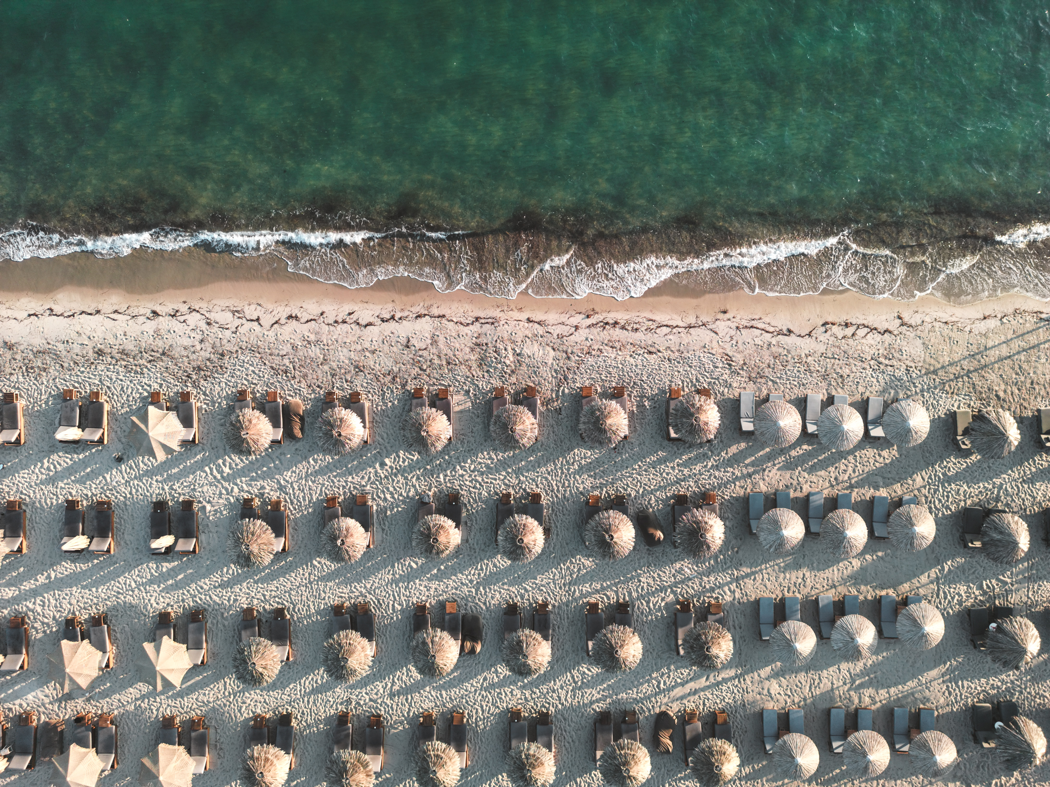 Luftaufnahme eines Strandes mit Reihen von Sonnenschirmen und Liegestühlen am Rand des türkisblauen Meeres.