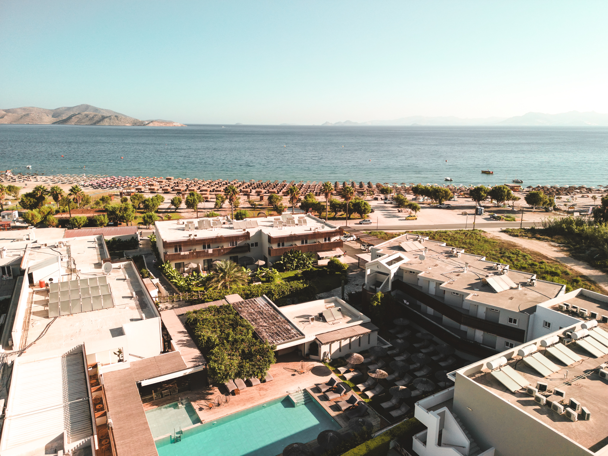 Luftaufnahme eines Resorts auf Kos mit Pool, grünen Palmen, Sandstrand voller Sonnenschirme und Blick auf das ruhige Meer mit Bergen im Hintergrund.