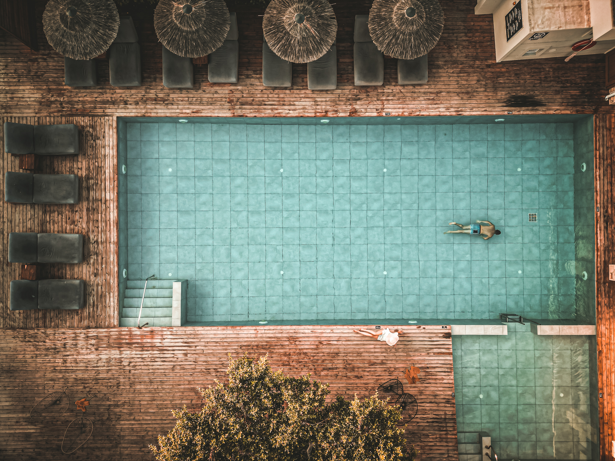 Draufsicht auf einen rechteckigen Pool mit türkisfarbenem Wasser, umgeben von Holzterrasse mit Sonnenliegen und Schirmen; eine einzelne Person schwimmt im Wasser.