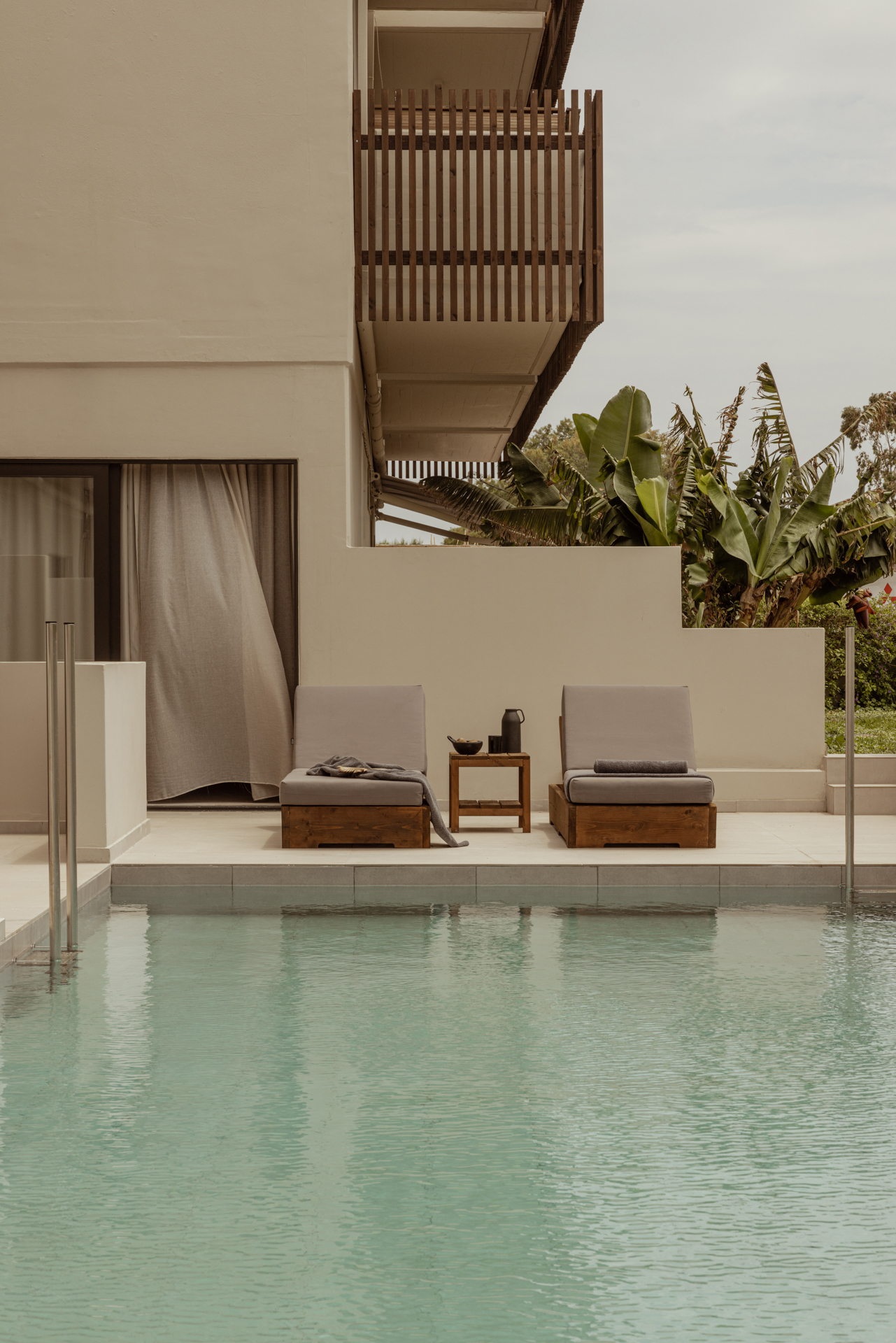 Moderne Poolterrasse mit zwei Liegen und einem kleinen Tisch vor einer Hotel-Suite, umgeben von tropischen Pflanzen.