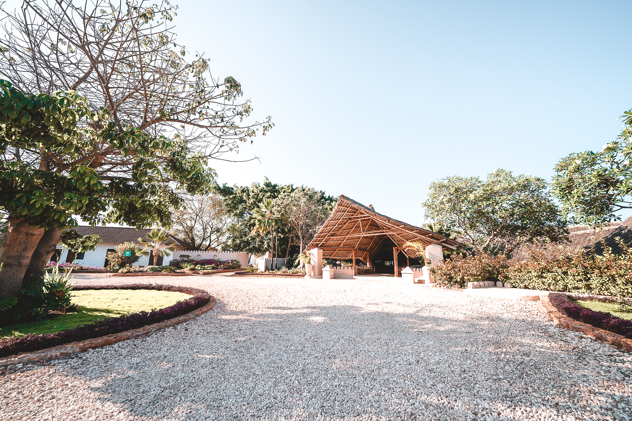 Einfahrt zum Resort mit einem offenen Empfangsgebäude mit Strohdach, umgeben von Bäumen und gepflegter Gartenanlage.
