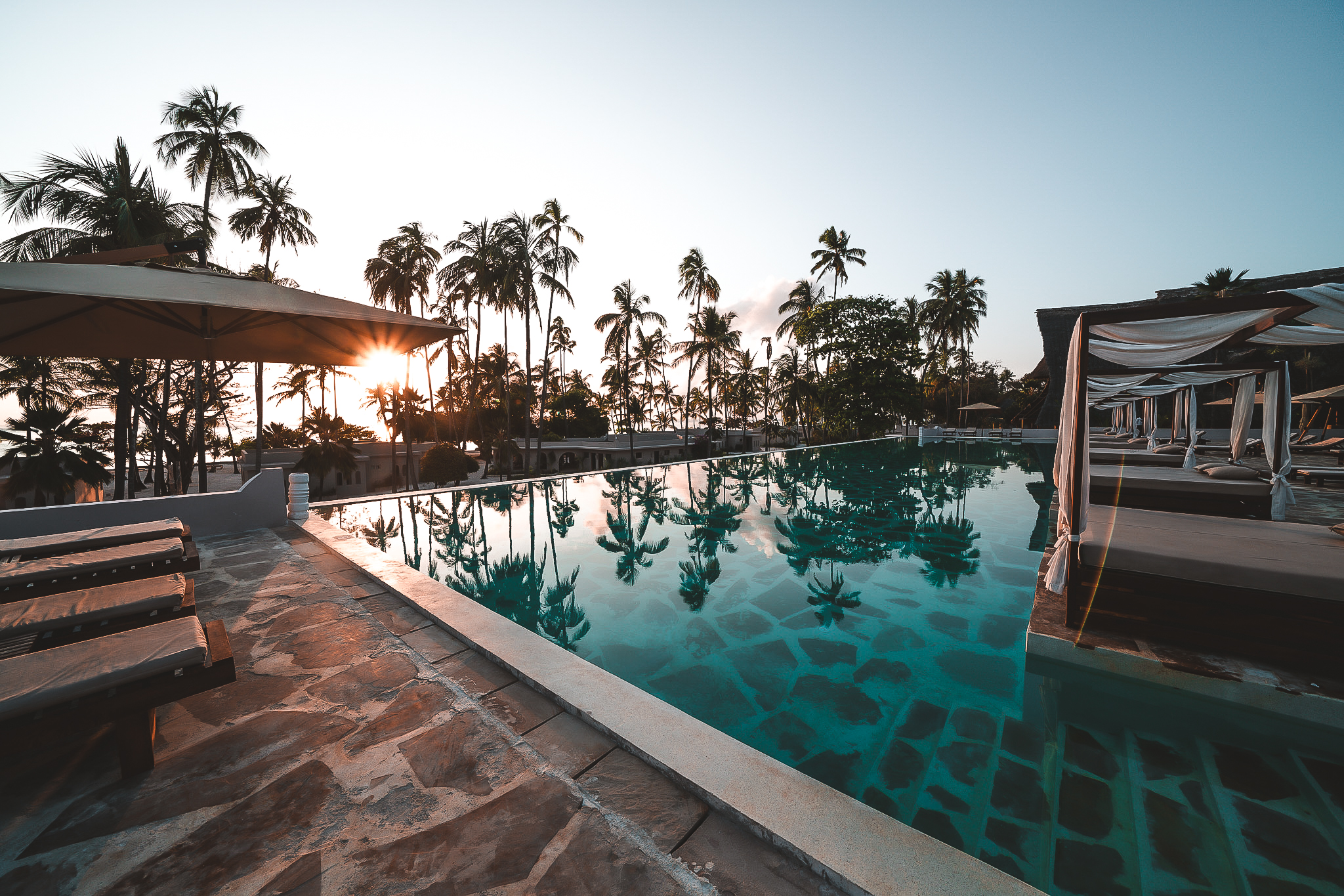Infinity-Pool im Diamonds Mapenzi Zanzibar bei Sonnenuntergang, gesäumt von Palmen, Sonnenliegen und balinesischen Betten.