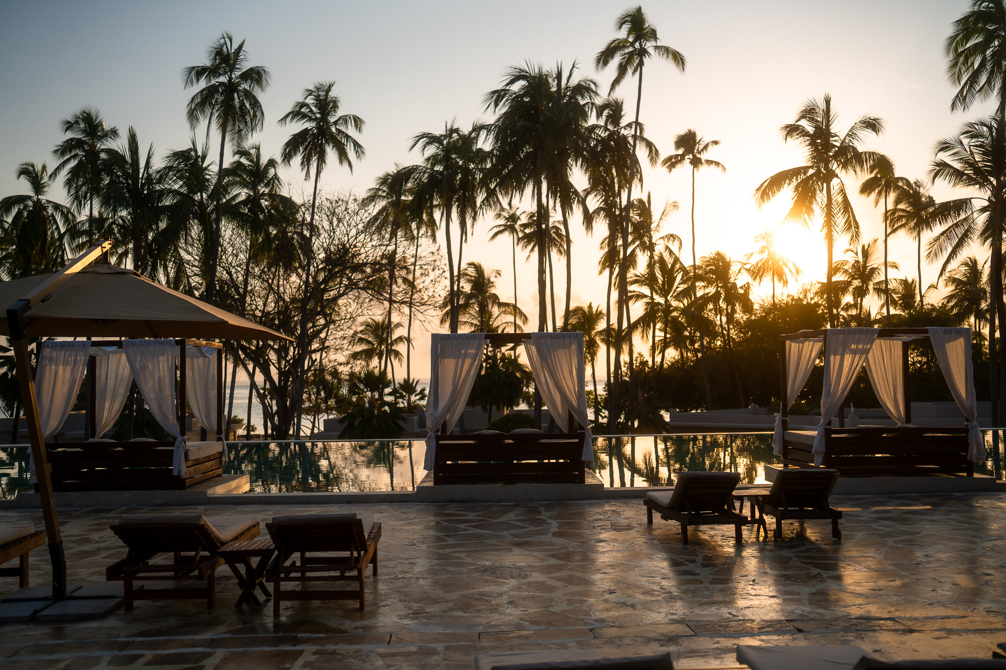 Romantische Daybeds mit weißen Vorhängen am Pool, umgeben von Palmen, während die Sonne malerisch im Hintergrund untergeht.