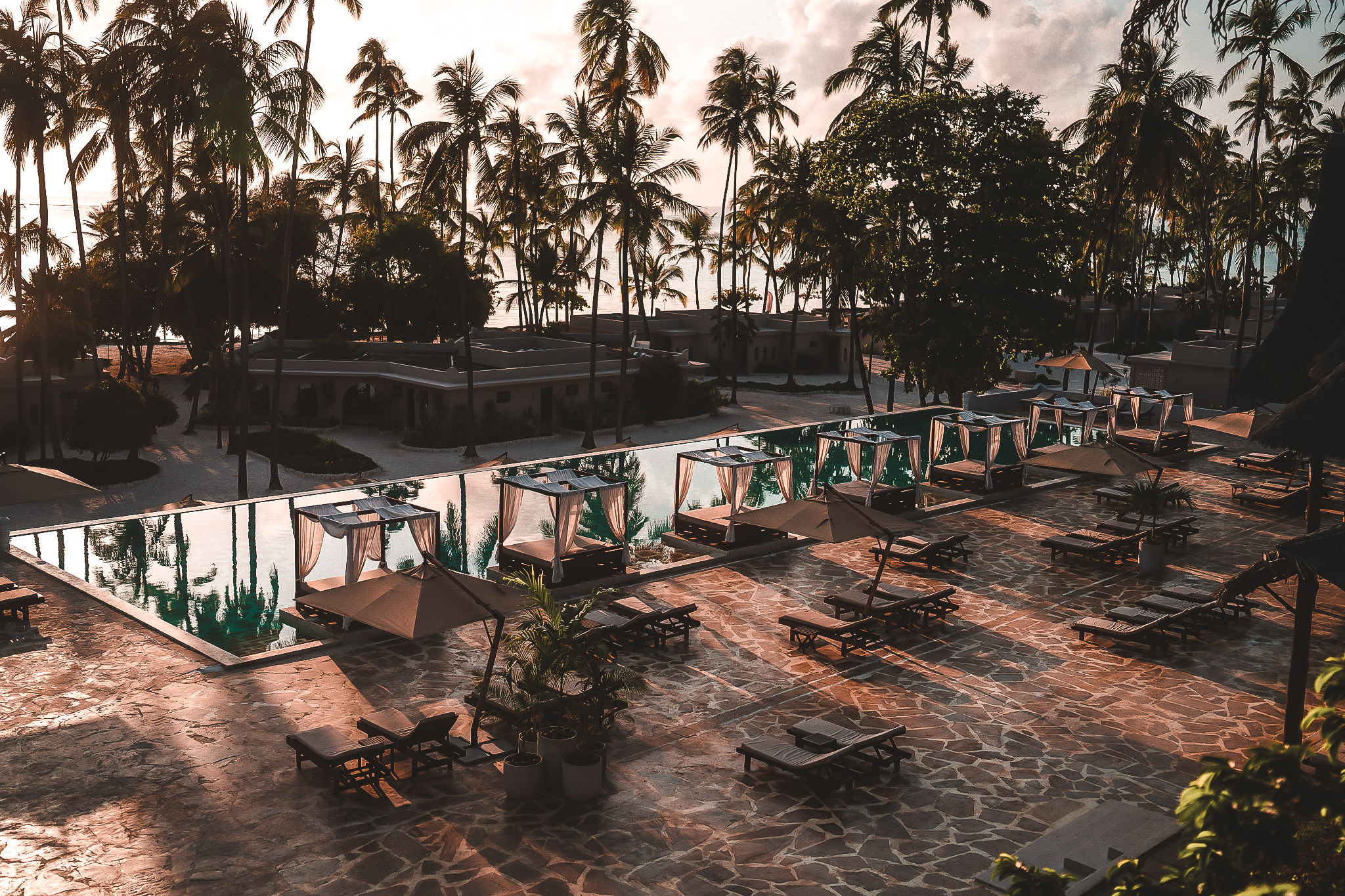 Poolbereich des Diamonds Mapenzi Beach Zanzibar bei Sonnenuntergang, mit Liegen, Sonnenschirmen und Palmen im Hintergrund.