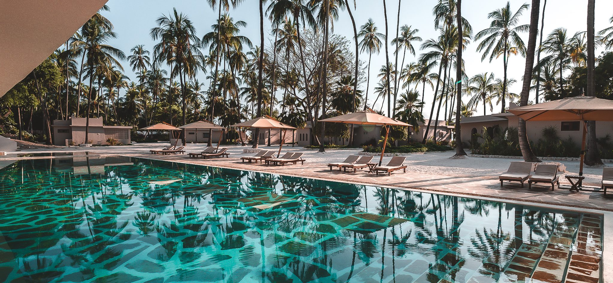 Großer türkisfarbener Pool mit Steinmuster, umgeben von Sonnenliegen und Sonnenschirmen, eingerahmt von Palmen und Strandvillen.
