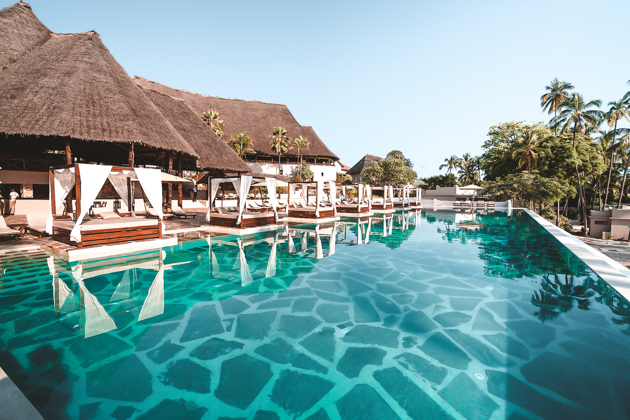 Luftaufnahme eines Strandresorts mit Infinity-Pool im Vordergrund, weißen Sandwegen und modernen Villen zwischen hohen Palmen, direkt am türkisfarbenen Meer gelegen.“ Alt-Text: „Luftaufnahme eines Strandresorts mit Infinity-Pool im Vordergrund, weißen Sandwegen und modernen Villen zwischen hohen Palmen, direkt am türkisfarbenen Meer gelegen.