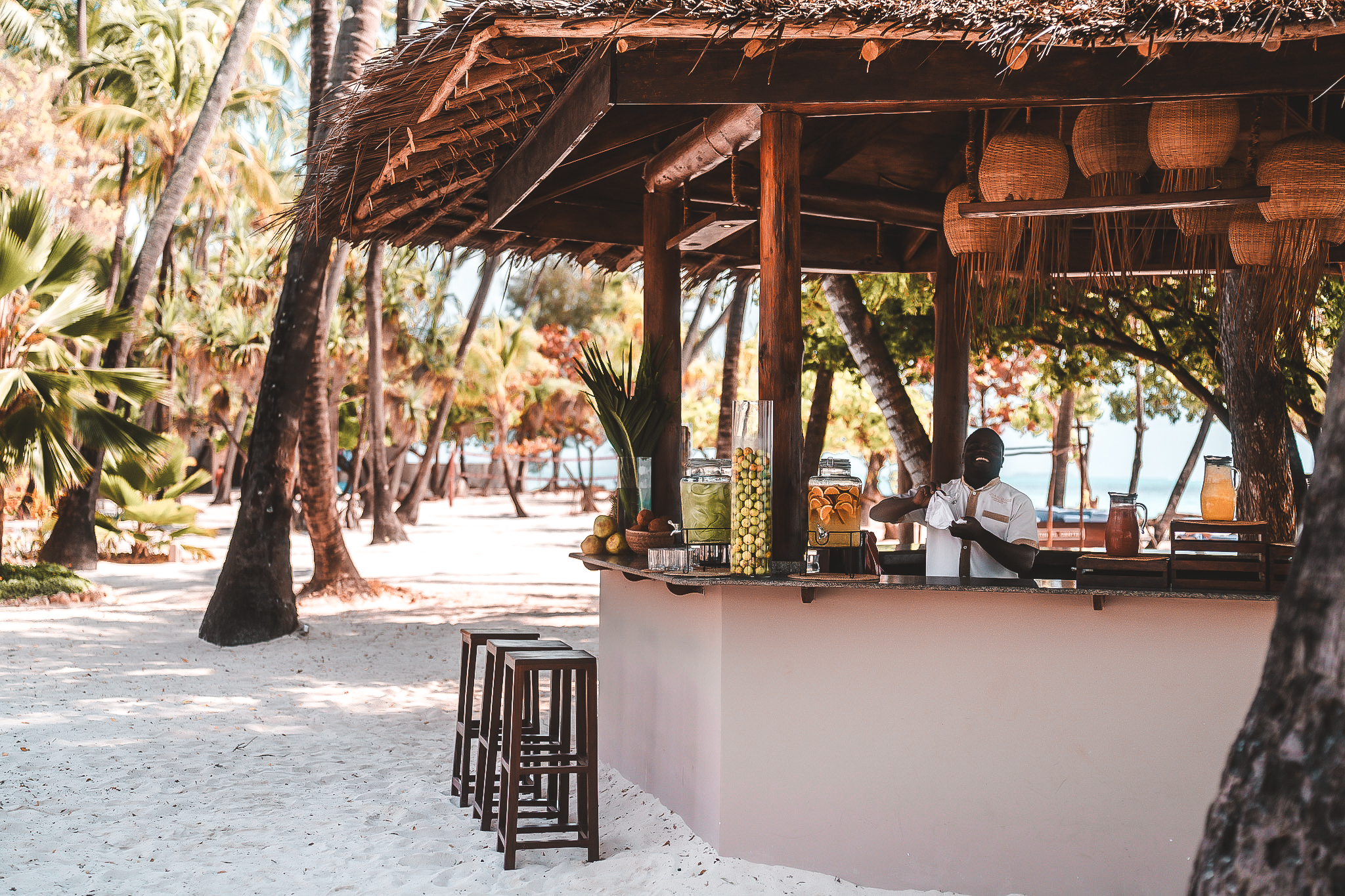 Offene Strandbar mit Strohdach, frischen Säften und gemütlichen Barhockern unter Palmen direkt am Sandstrand.