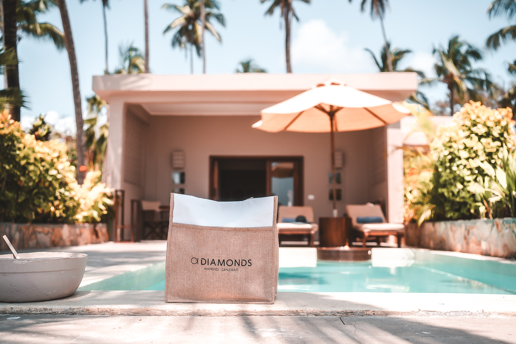 Blick auf eine private Villa mit Pool im Resort Diamonds Mapenzi Zanzibar, im Vordergrund eine Strandtasche mit Logo, dahinter Liegestühle und Sonnenschirm zwischen tropischer Vegetation.