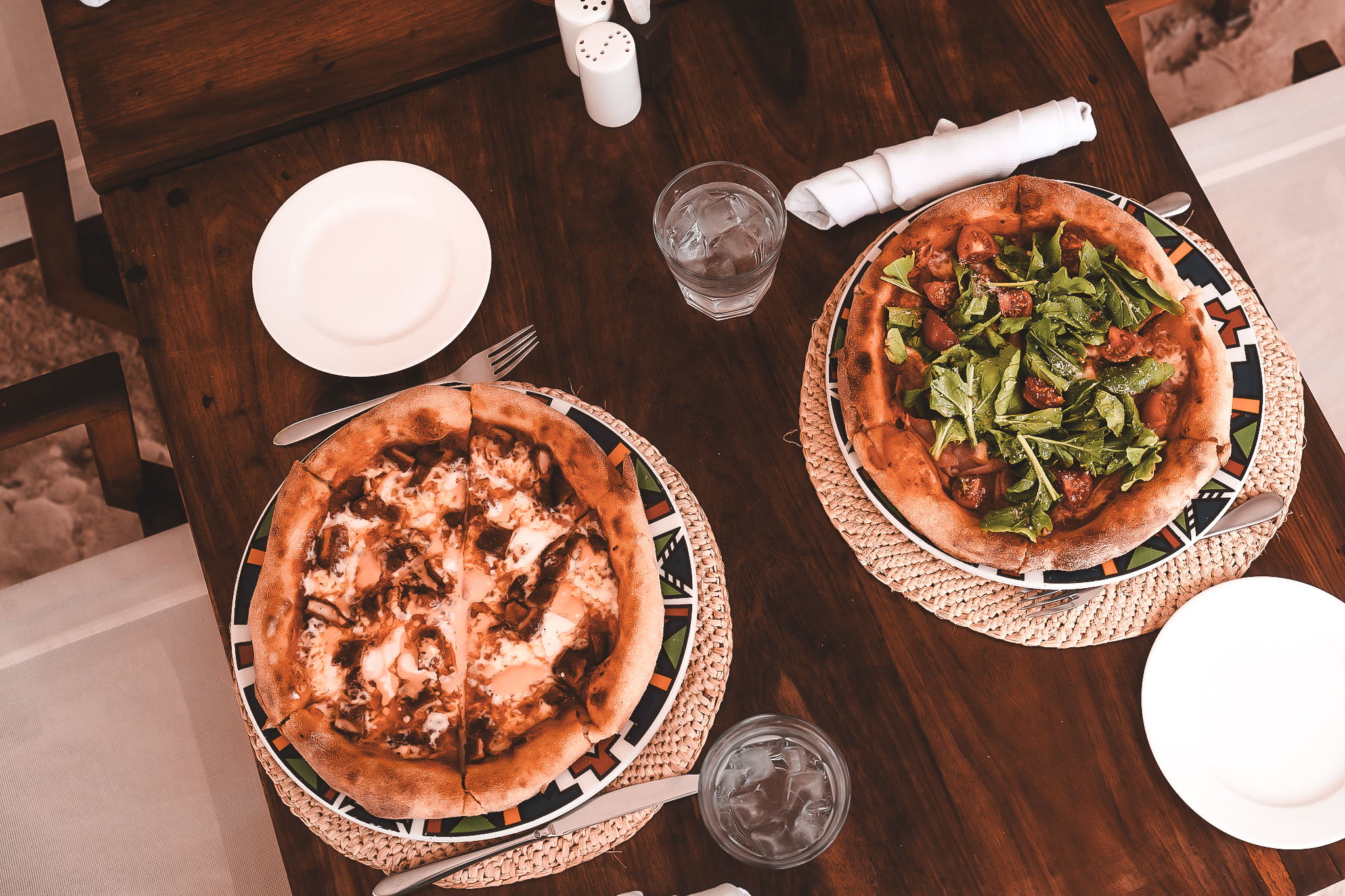 Frisch gebackene Pizzen – eine mit Käse und Pilzen, die andere mit Rucola und Cherrytomaten – serviert auf bunten Tellern am Holztisch.