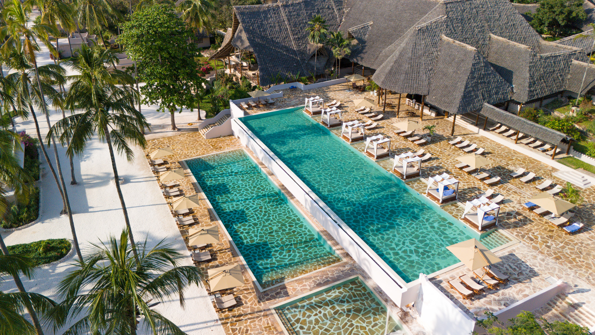 Weitläufige Poolanlage mit eleganten Liegen und Pavillons, umgeben von Palmen und traditioneller Architektur.